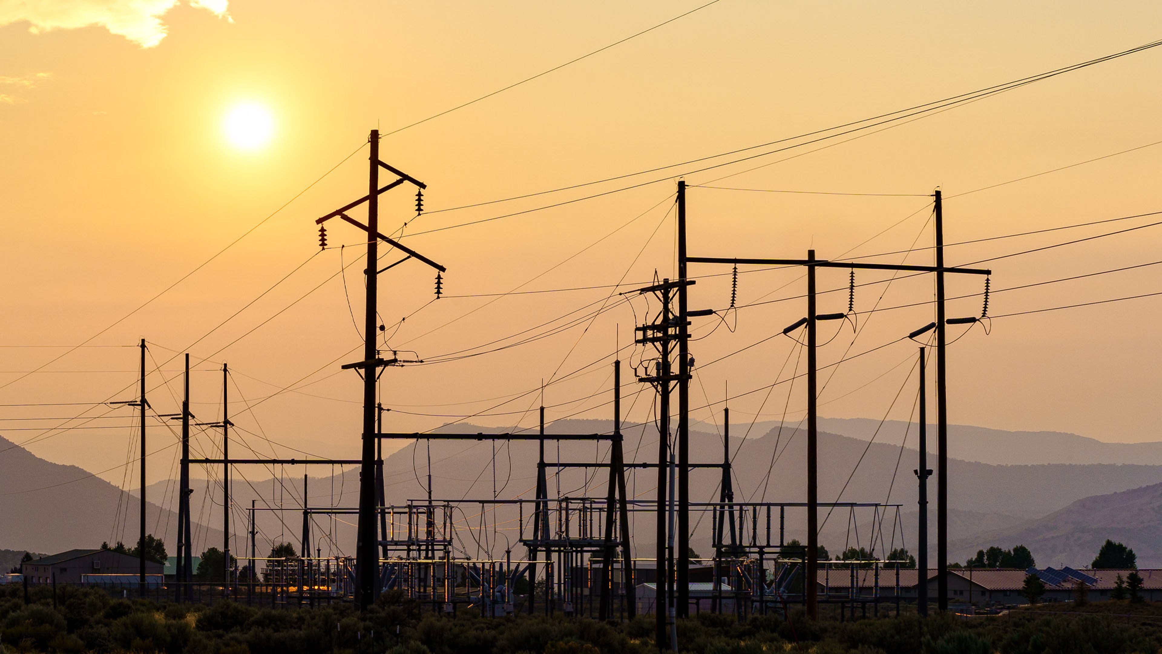 Utility poles and power lines silhouetted against a golden sunset with mountains in the background.