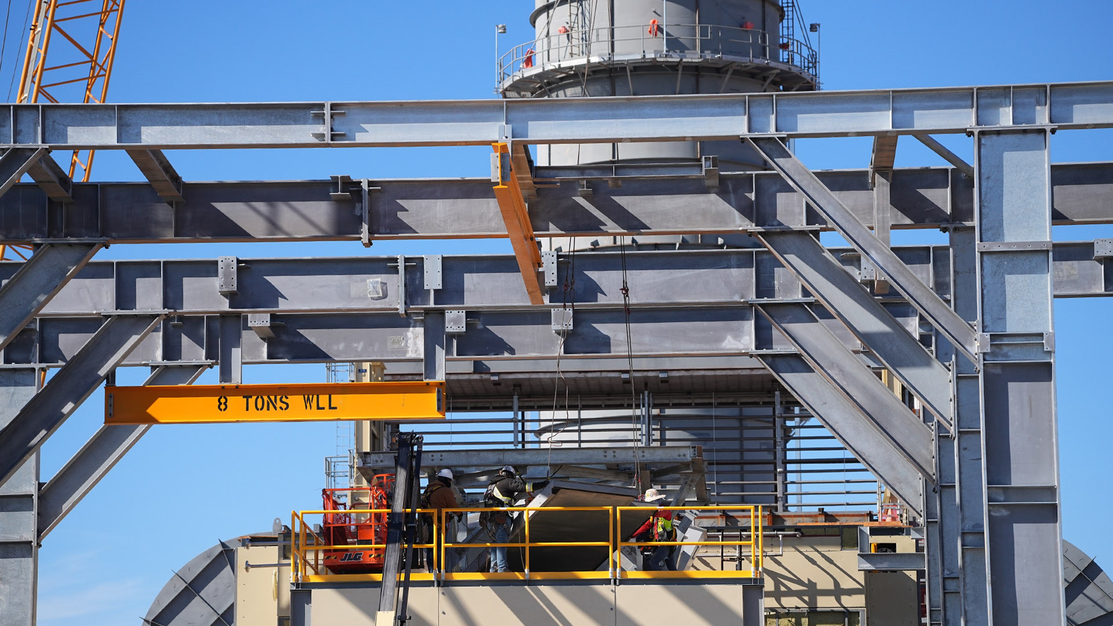 Workers on a platform within a steel-framed construction site under a blue sky.