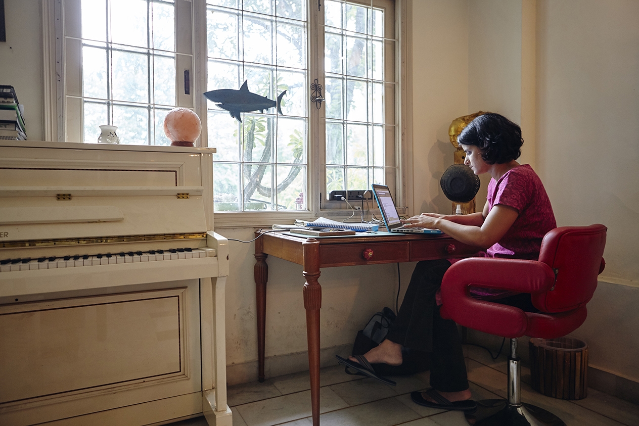 Женщина сидит за столом у окна и работает на ноутбуке.
