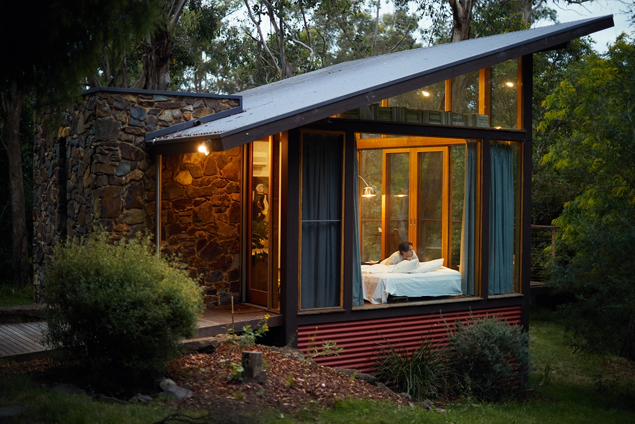 Una casa nel bosco con vetrate a tutta altezza e una persona che si rilassa all'interno.