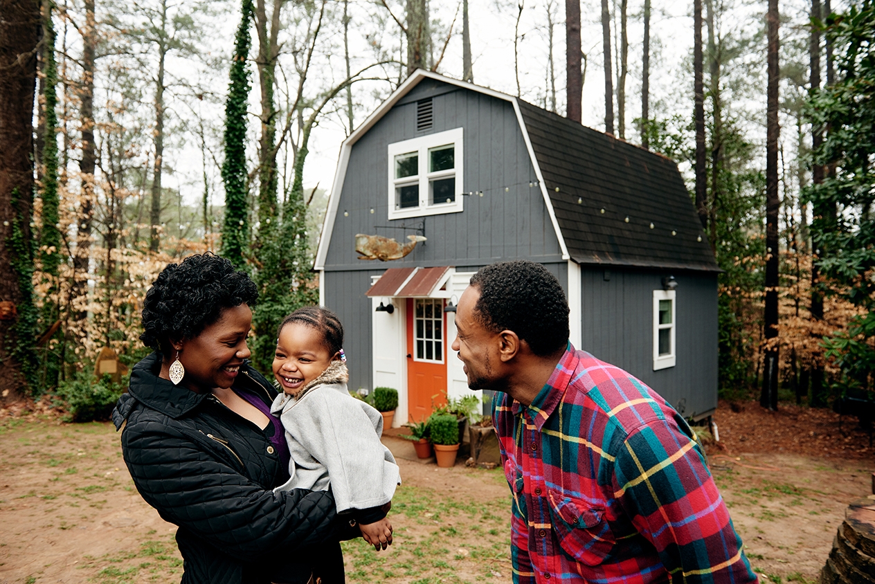 Two adults and a child, laughing, outside a barn-shaped house in the forest.
