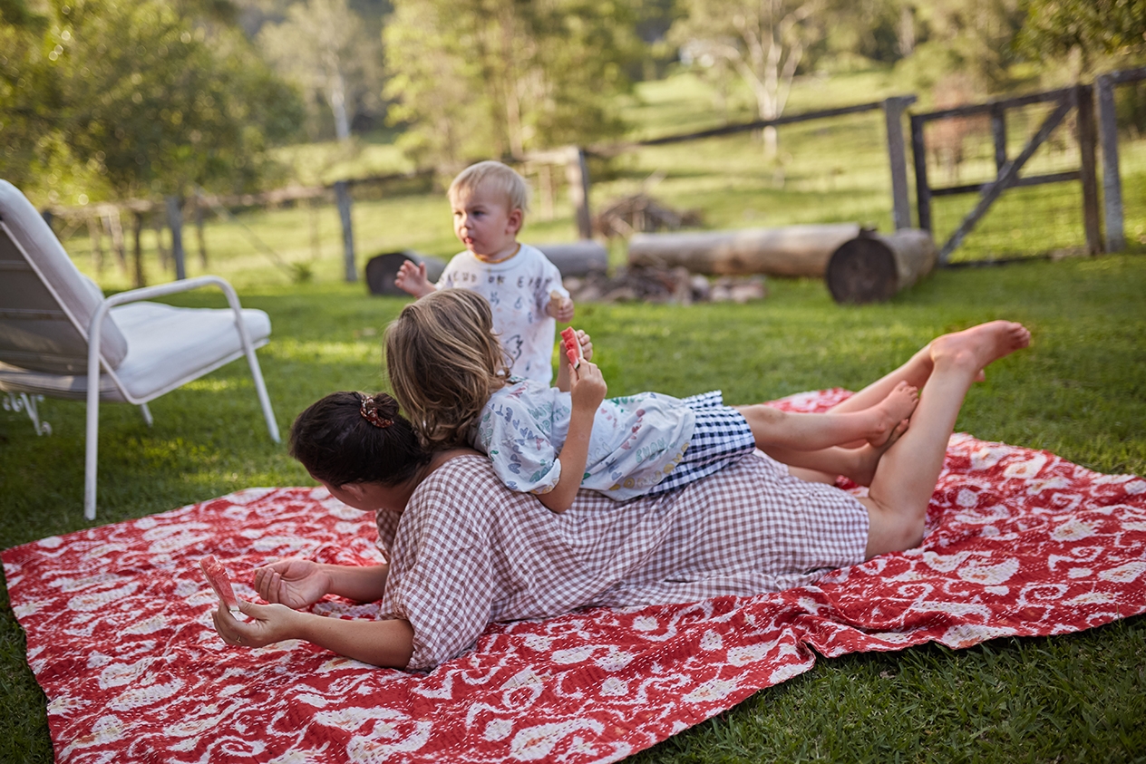 Una persona adulta y dos niños están tumbados en una manta del colores sobre el césped, al aire libre.