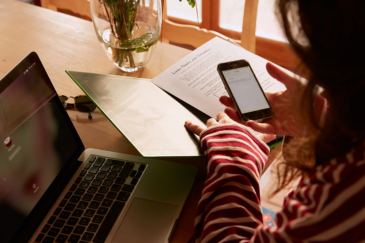 Eine Person prüft Informationen auf ihrem Laptop und ihrem Mobiltelefon, während sie auf gedruckte Dokumente verweist.