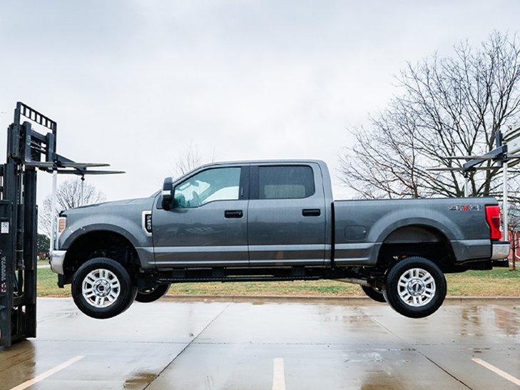 a pickup truck is hanging between two forklifts to demonstrate the strength of Pella's fiberglass
