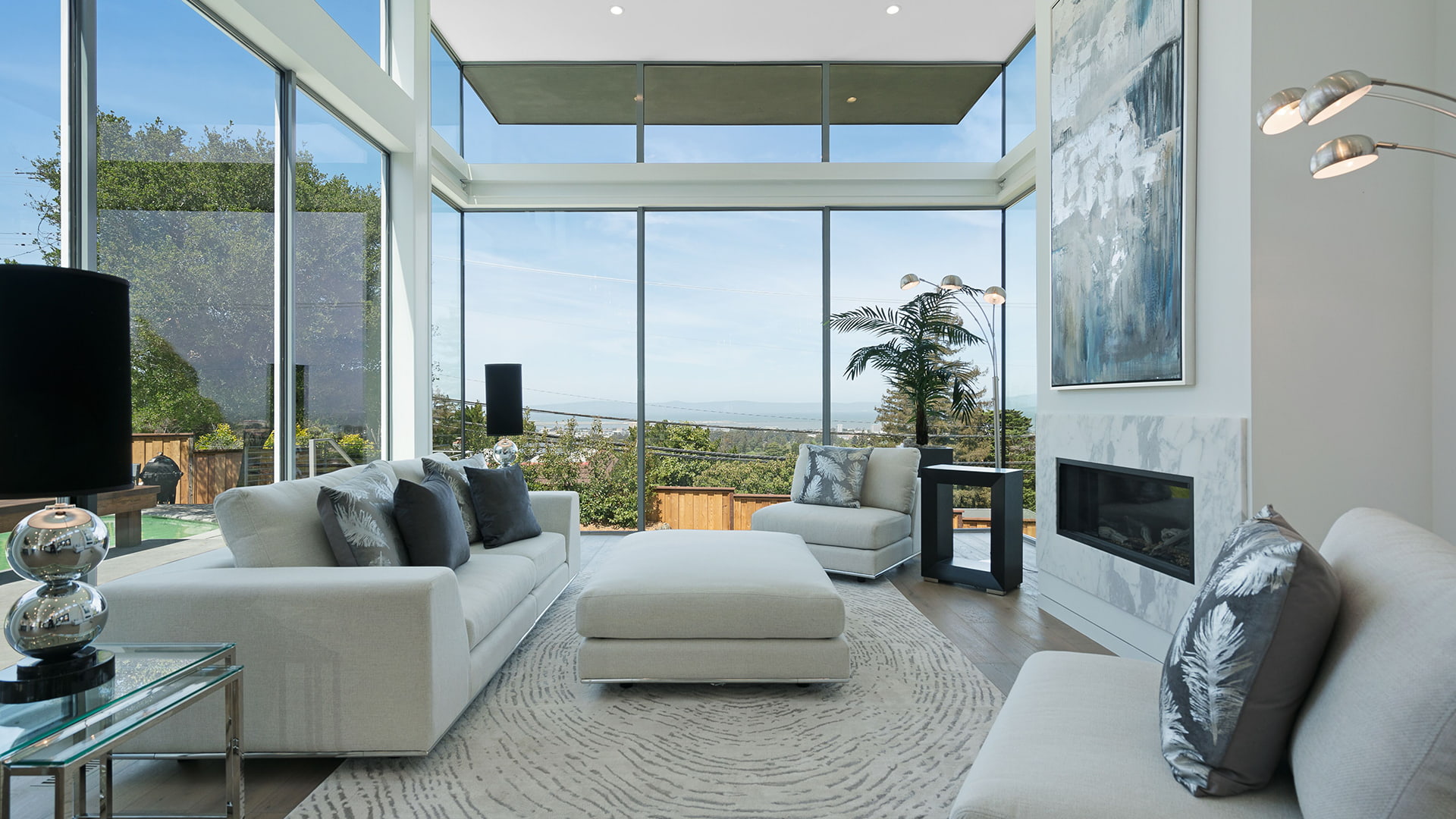 Window walls surround a living room with a white sofa and several accent pillows
