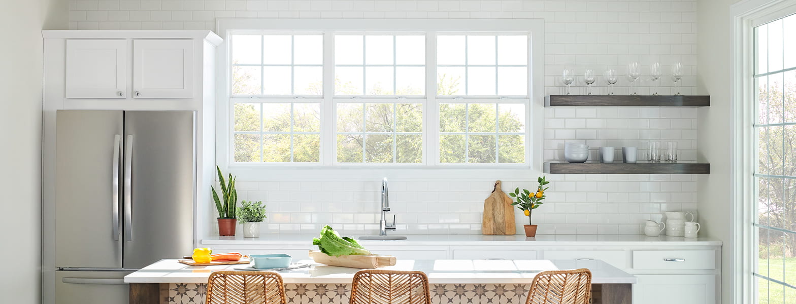 Three vinyl double-hung windows between a fridge and shelves above a white kitchen counter.