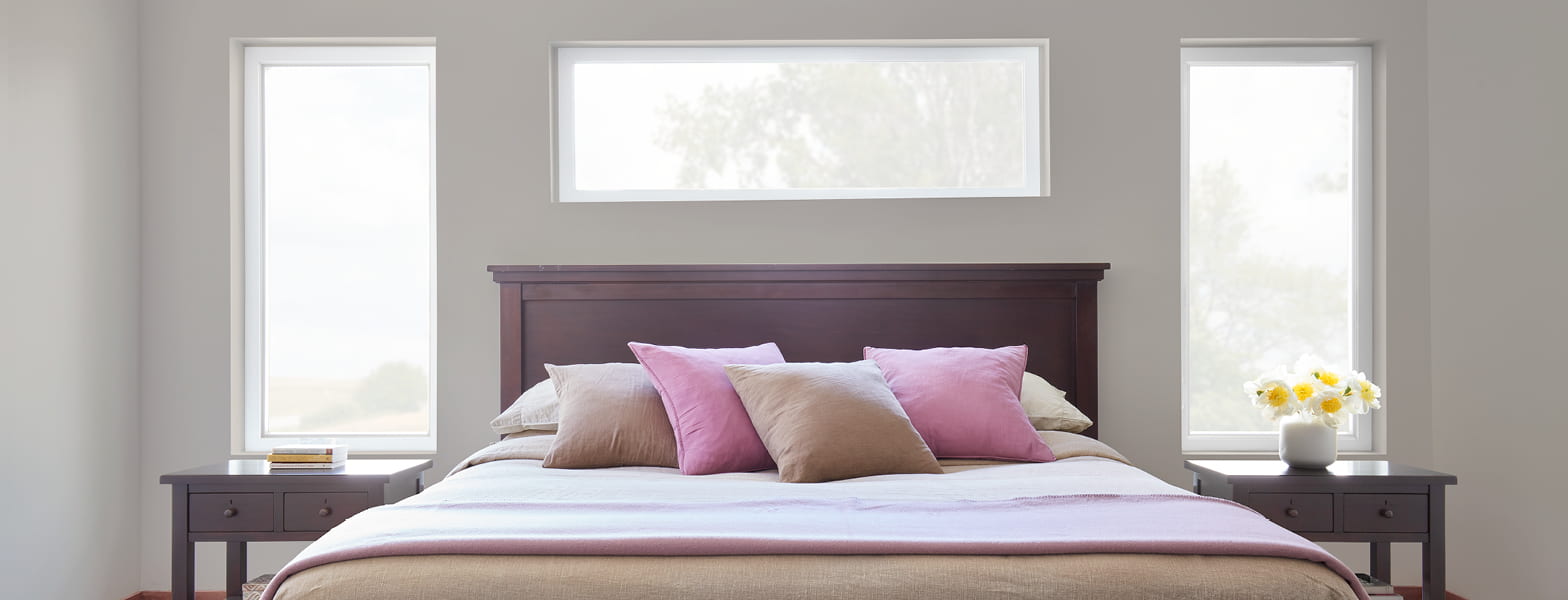 a large bed with pink and tan pillows below and in between Pella vinyl windows