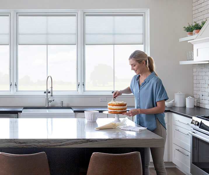 A woman is icing a cake on a large island and three large casement windows are behind her.