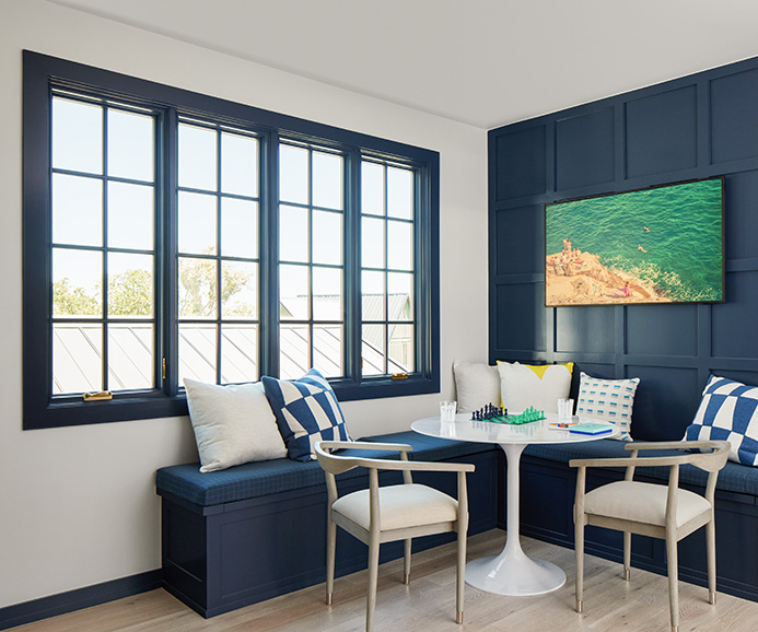 A woman is icing a cake on a large island and three large casement windows are behind her.