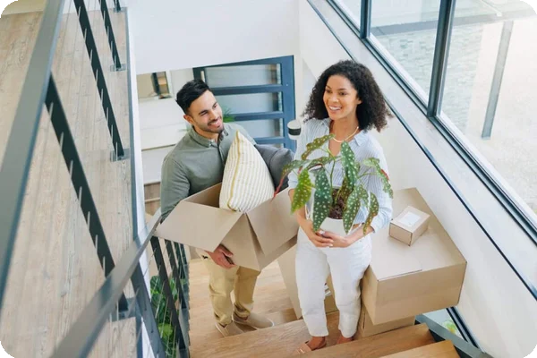 a couple happily walks up the stairs carrying boxes and plants to move into their new apartment