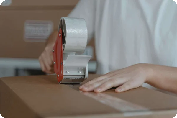 Person's hands apply clear tape to seal a brown cardboard box with a tape dispenser.