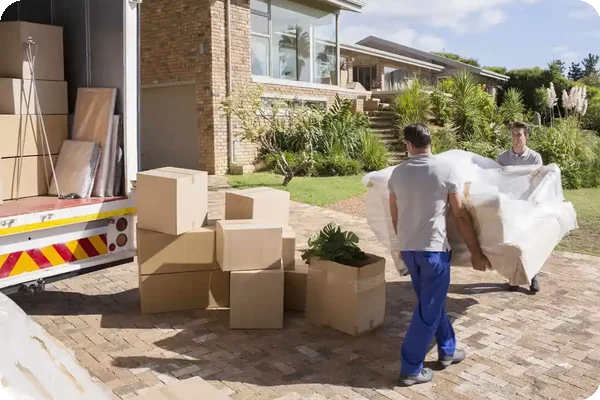 Moving workers carrying a wrapped couch from a home to a moving truck with boxes stacked nearby