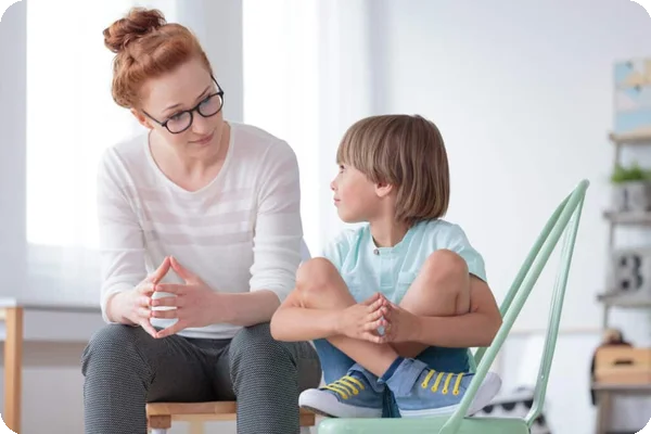 Red-haired woman in glasses looks at a boy with crossed legs on a green chair.