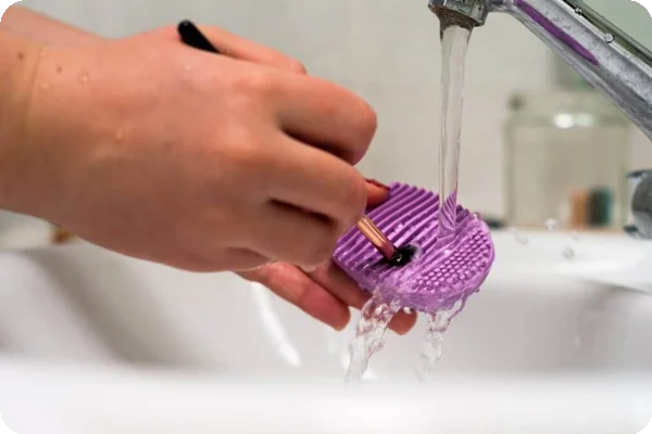 someone rubs a makeup brush on a cleaning tool underneath a stream of running water from a skink faucet