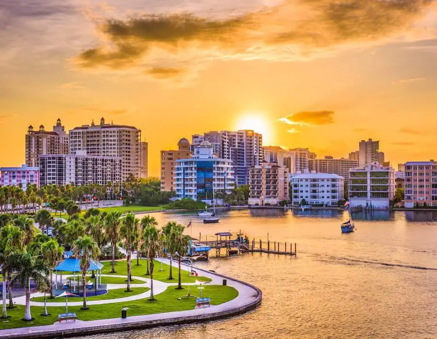 Sunset view of Sarasota, Florida, with waterfront buildings, a small marina, and palm trees in the foreground.