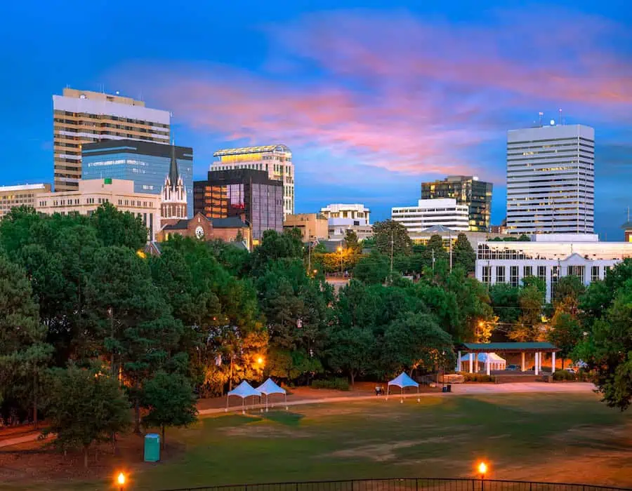 Skyline of Columbia, South Carolina, with mid-rise buildings and a tree-filled park in the foreground at sunset.