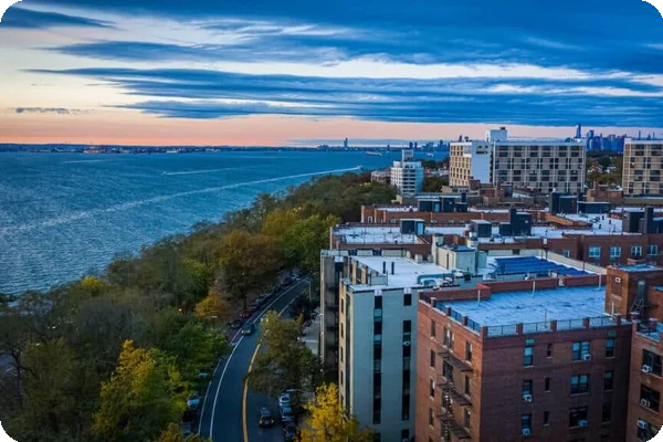Elevated view of a cityscape with apartment buildings, a winding road, a bay, and a distant skyline at dusk.