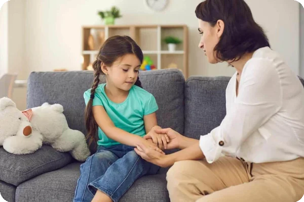 A mother holds her daughter's hands and speaks to her while sitting on a couch.
