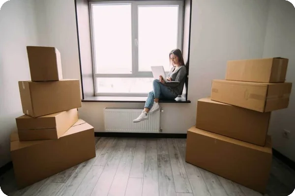 A young woman sits on a windowsill with a laptop. There are a few cardboard boxes stacked around her.