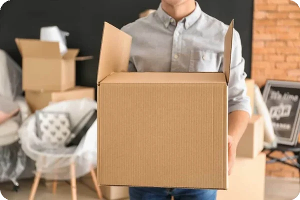 A man carrying a cardboard box. In the background, there is a chair wrapped with shrink wrap and several more boxes
