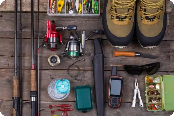 Assorted fishing equipment including rods, reels, lures, boots, and tools arranged on a wooden surface.