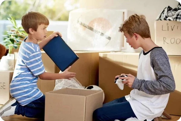 Two young boys pack their belongings into a cardboard box. One holds a video game controller
