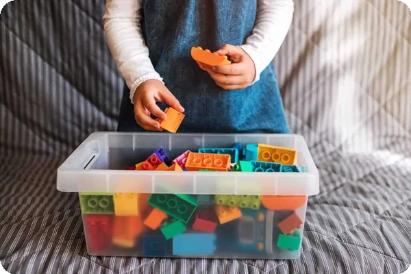 A young girl puts colorful lego blocks in varying sizes into a clear plastic tub