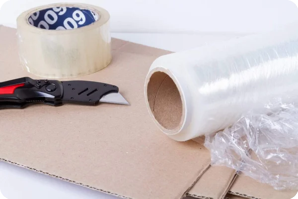 A box cutter, tape, shrink wrap, and cardboard sit on a table