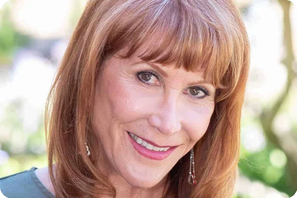 Smiling woman with reddish-brown hair and bangs, wearing makeup and earrings, looking at the camera.