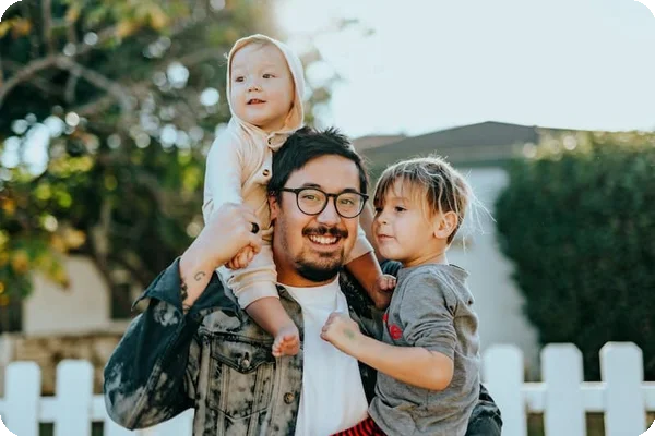 A smiling man carries two young children, one on his shoulders, one on his hip, outdoors by a fence.