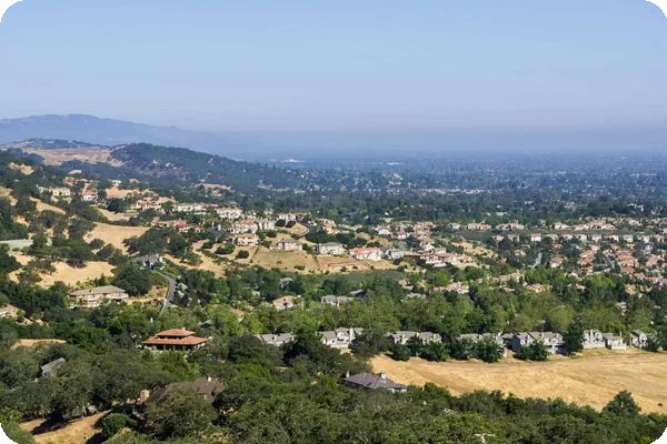 Hilly suburban landscape with houses, trees, and dry hills overlooking a hazy distant city.