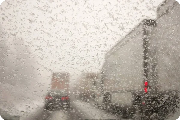 Cars and trucks driving on snow packed roads taken through a windshield covered with snowflakes.