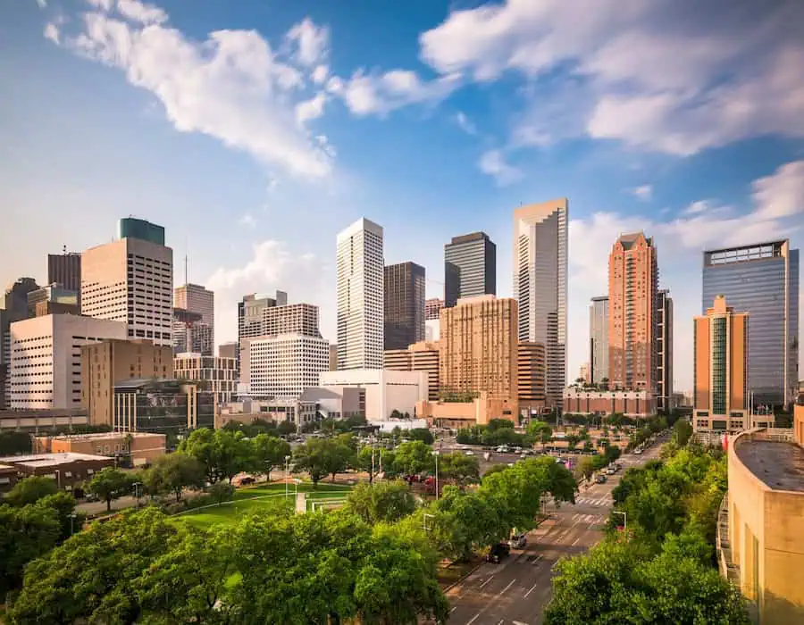 Modern skyline of Houston, Texas, featuring tall office buildings and tree-lined streets under a partly cloudy sky.