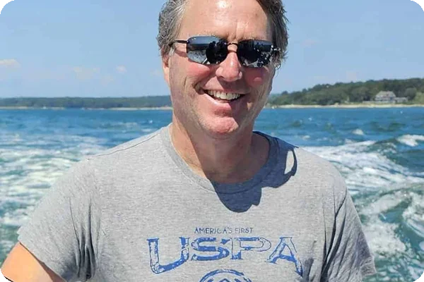 Smiling man in sunglasses and grey USPA t-shirt on a boat with water and land in the background.