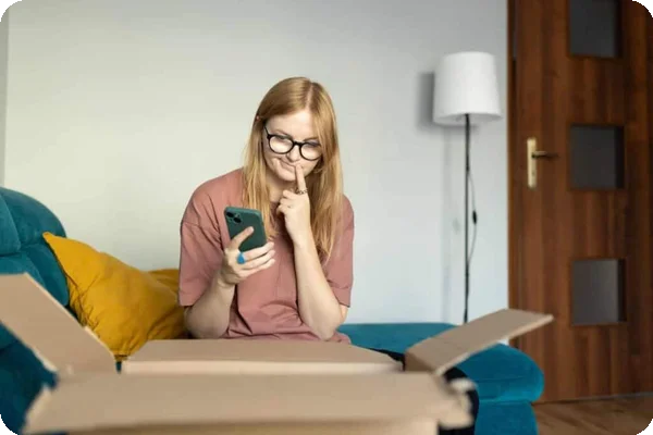 A woman sits on a chair in front of an open moving box. She has a contemplative expression and is holding her phone in one hand.