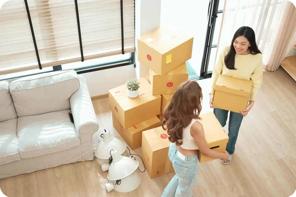 Two women carrying boxes into a home. There is a pile of more boxes next to them.