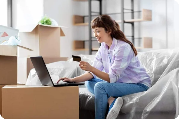 A woman sits on a covered couch holding a credit card in front of a laptop. She's surrounded by moving boxes.