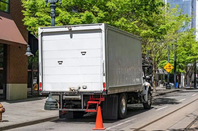 A moving truck parked along a sidewalk has orange traffic cones around it 