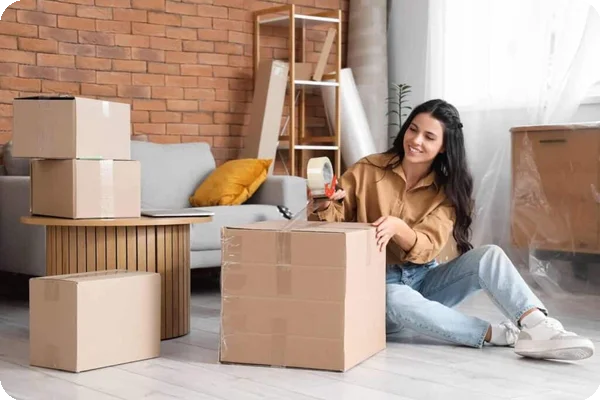 A woman sitting on the floor tapes a cardboard box closed. There are others already packed beside her