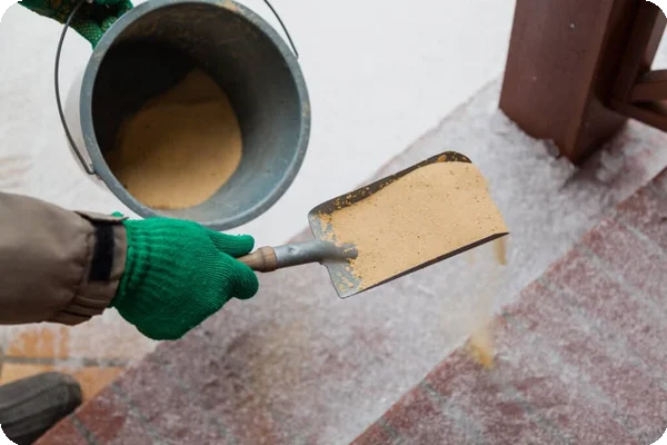 Gloved hand shoveling sand from a bucket onto an icy, brick-paved surface.