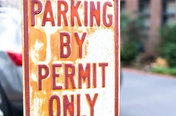 A white sign with red lettering that reads Parking By Permit Only