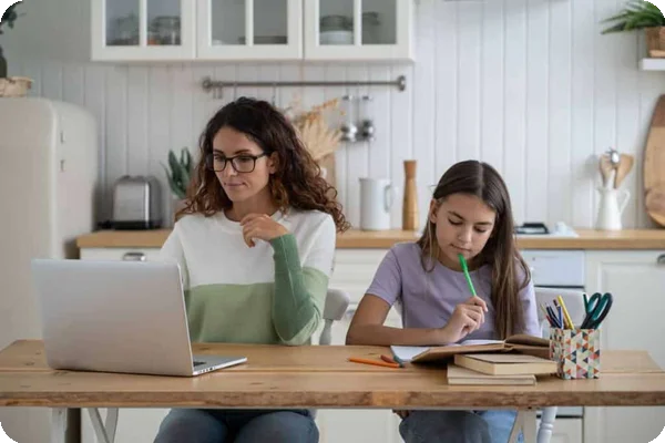 A mother works on a laptop next to her child at a kitchen table