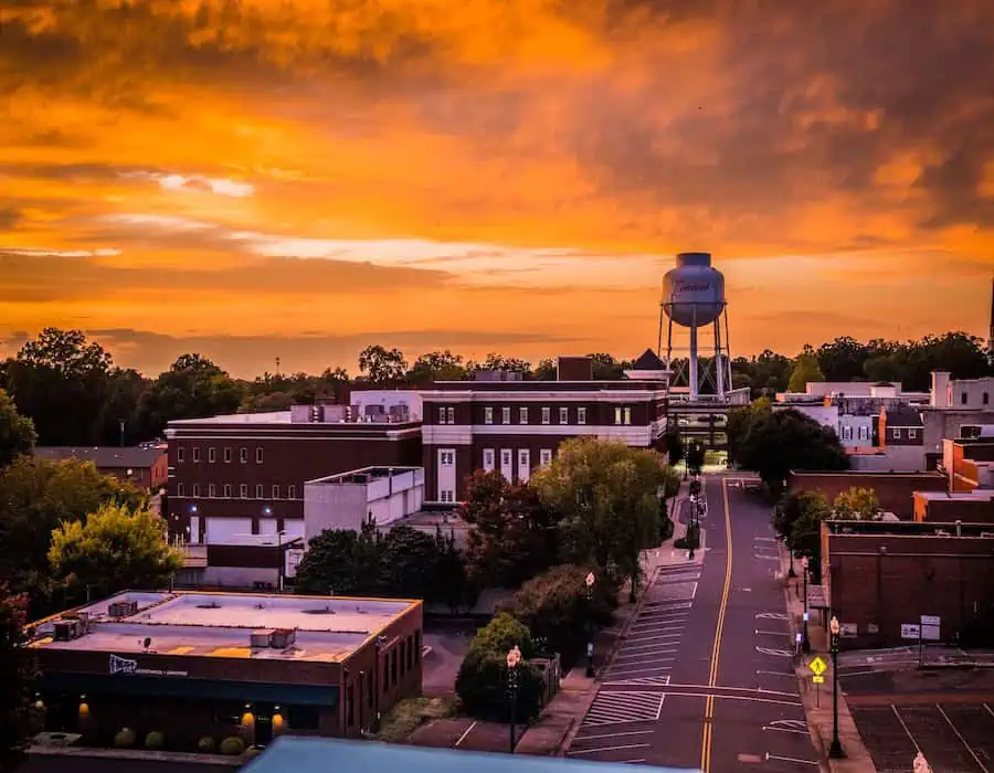Main street in Concord, North Carolina, with low-rise buildings, trees, and a water tower in the background during a vivid orange sunset.