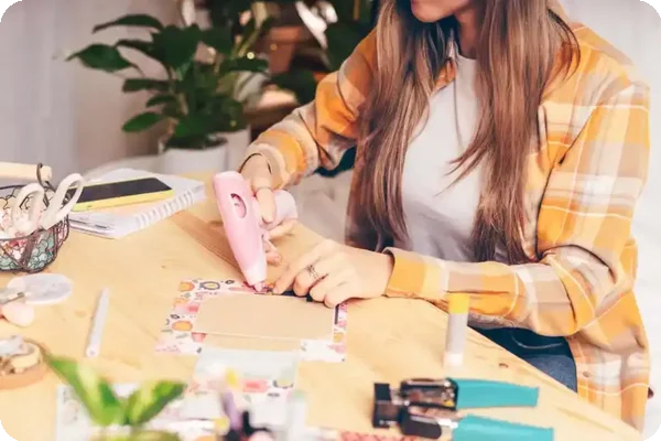Woman working on a scrapbook with a glue gun and decorative paper at home.