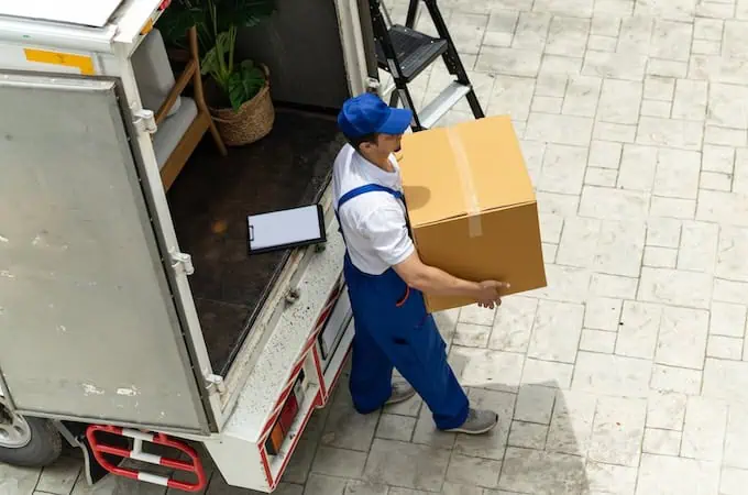 A mover in blue overalls carries a box from a moving truck on a sunny day