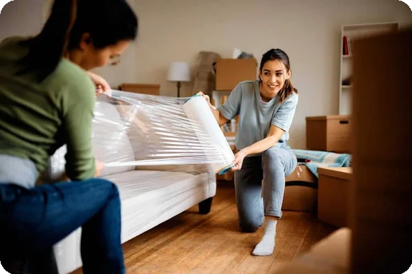 Two women use shrink wrap on a couch