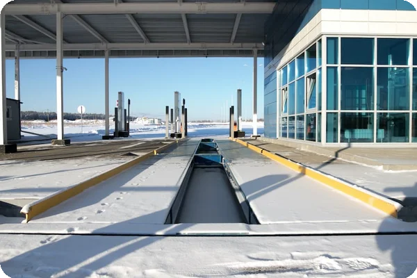 Snowy vehicle inspection lanes at a modern weigh station with an overhead canopy and a glass-fronted building.