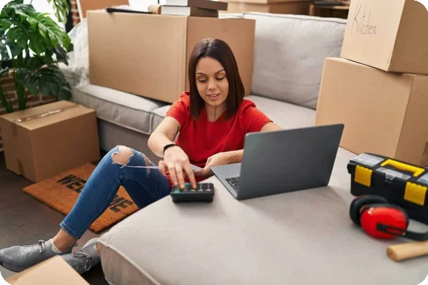 A woman sits on the floor and inputs something on a calculator and her laptop. There are moving boxes around her