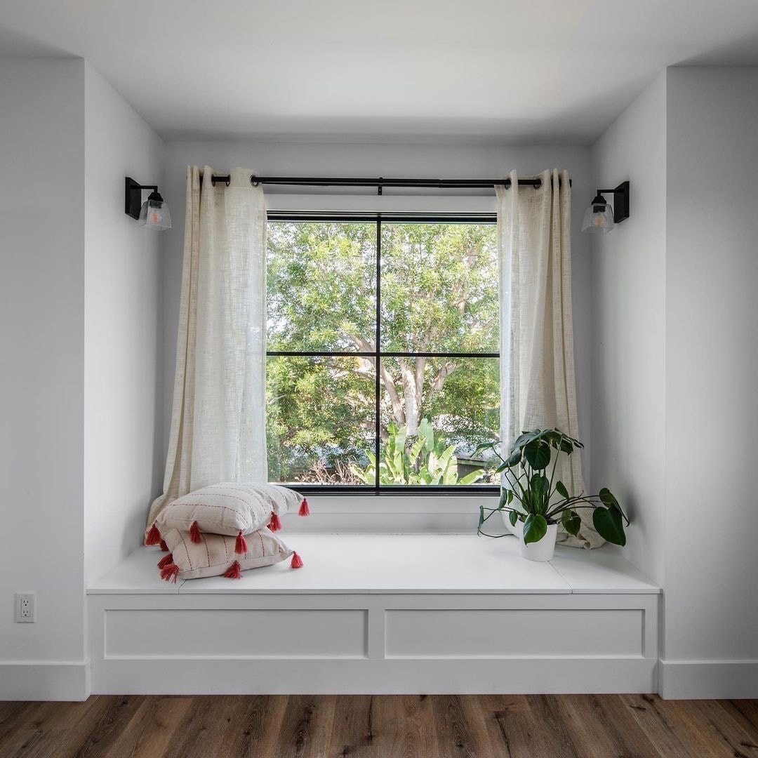 A black picture window decorated with curtains sits above a window seat.