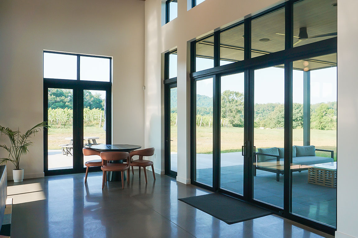 Glass walls consisting of black hinged doors and fixed windows frame the corner of a winery great room.